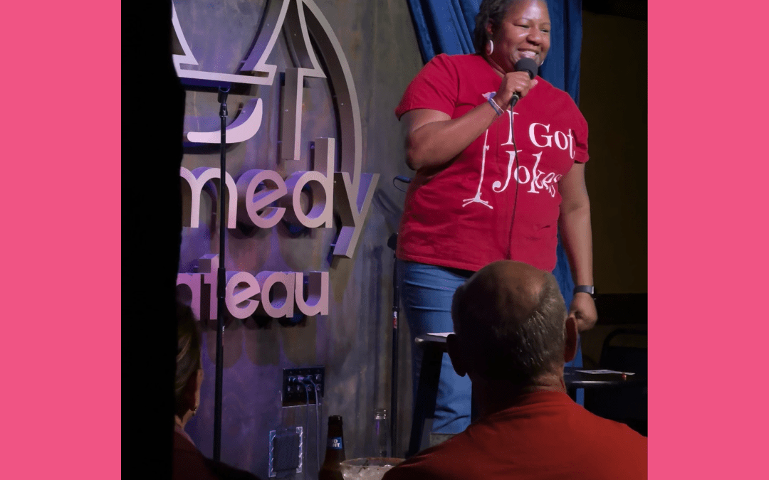 black woman standing on stage holding a microphone and wearing a t-shirt that says "I Got Jokes" in white lettering