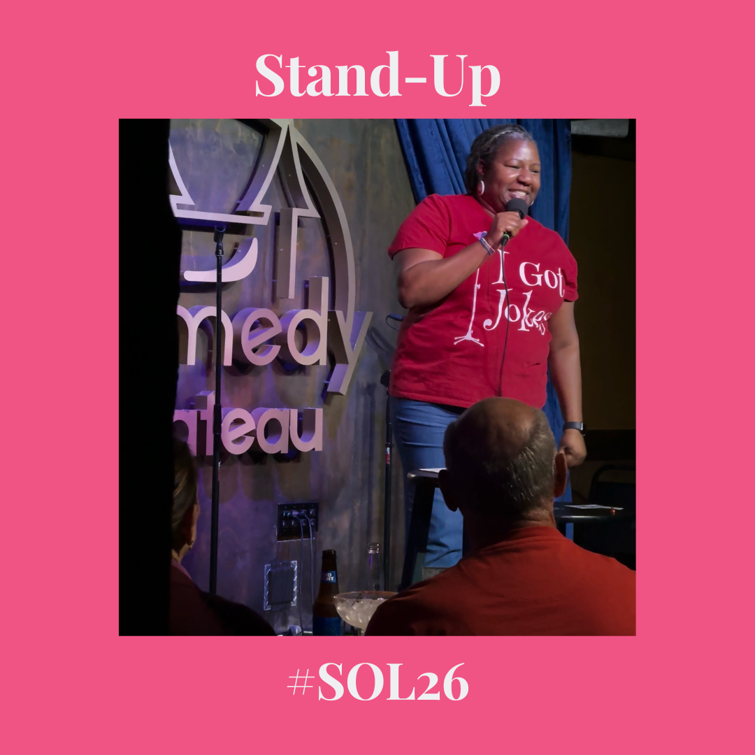 black woman standing on stage holding a microphone and wearing a t-shirt that says "I Got Jokes" in white lettering